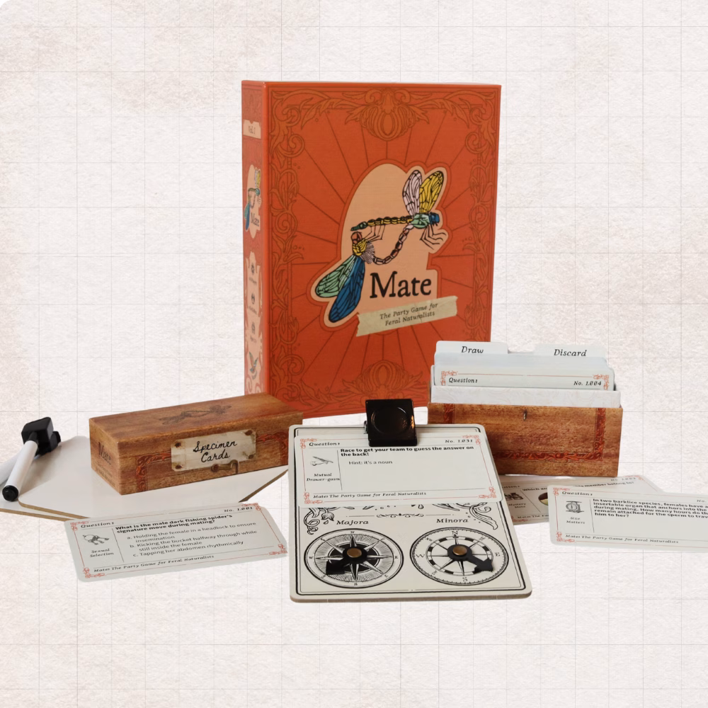In front of Mate: The Party Game for Feral Naturalists, the contents are laid out, including a specimen card box, clipboard with scoring compasses, and two drawing boards and markers.