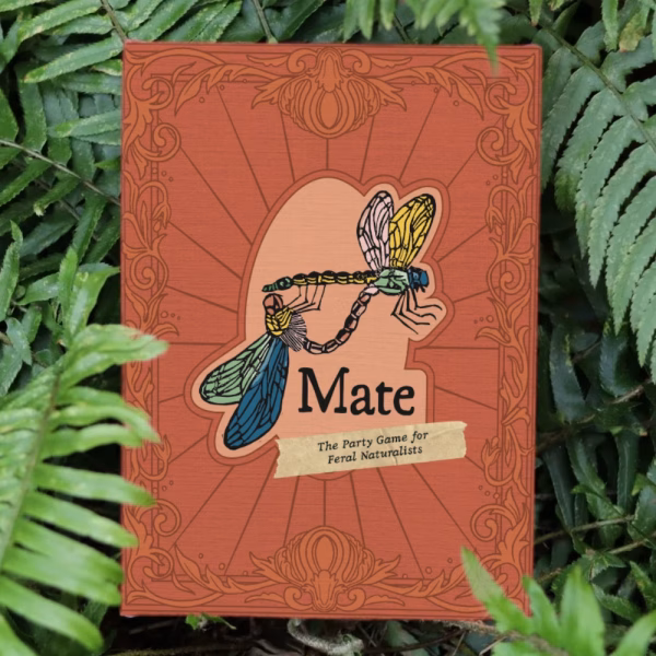 Mate: The Party Game for Feral Naturalists box in a bed of ferns. It has an ornate victorian banner and depicts two mating linocut dragonflies.