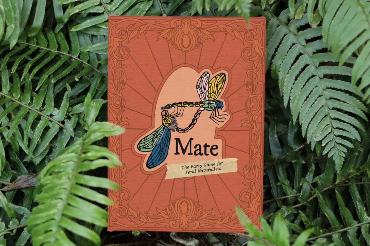 Mate: The Party Game for Feral Naturalists box in a bed of ferns. It has an ornate victorian banner and depicts two mating linocut dragonflies.